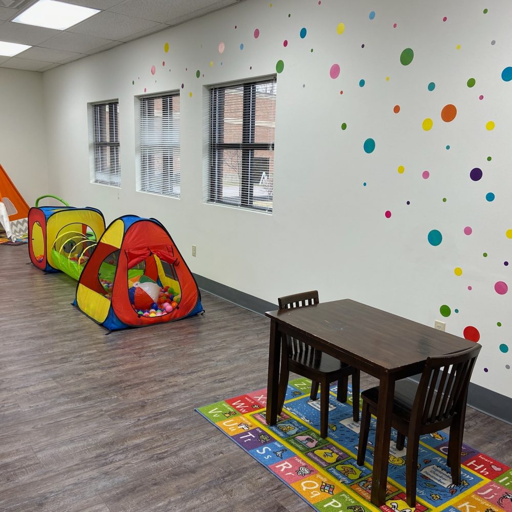 Brightly colored children's playroom with polka-dot walls, play tents, a ball pit, a wooden table, and alphabet rugs.