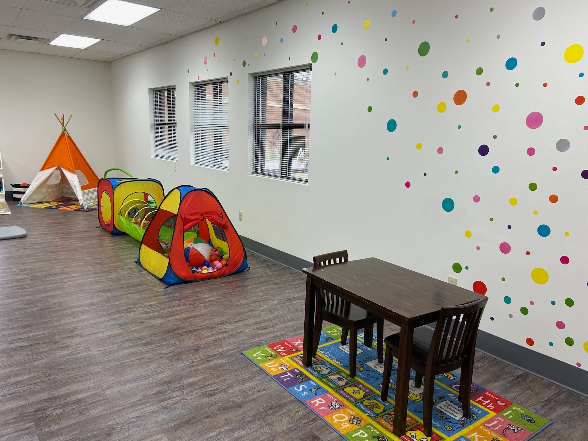Brightly colored children’s playroom with polka-dot walls, play tents, a ball pit, a wooden table, and alphabet rugs. Brightly colored children's playroom with polka-dot walls, play tents, a ball pit, a wooden table, and alphabet rugs.